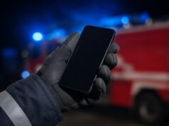 Kostenlose Notruf-App: Feuerwehrangehörige können per Knopfdruck Hilfe rufen Behandschuhte Hand einer Feuerwehrkraft hält ein Smartphone, im Hintergrund ein Löschfahrzeug mit Blaulicht.