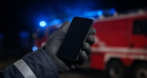 Kostenlose Notruf-App: Feuerwehrangehörige können per Knopfdruck Hilfe rufen Behandschuhte Hand einer Feuerwehrkraft hält ein Smartphone, im Hintergrund ein Löschfahrzeug mit Blaulicht.