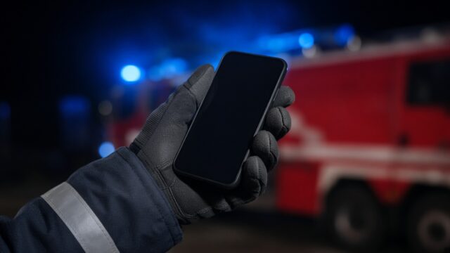 Behandschuhte Hand einer Feuerwehrkraft hält ein Smartphone, im Hintergrund ein Löschfahrzeug mit Blaulicht.