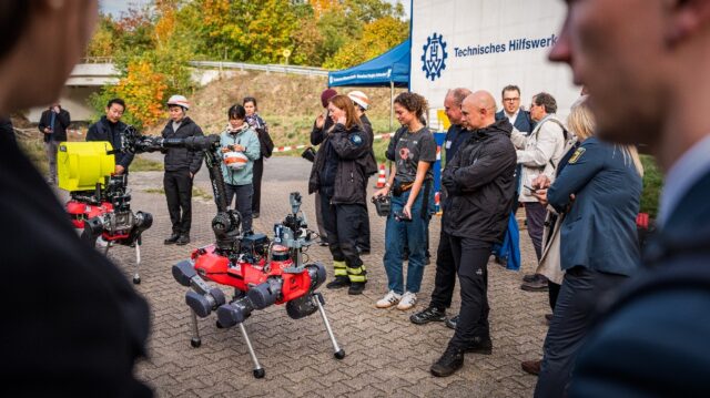 K1_thw_synergise_roboter_LeonKarl_930 Zwei rote vierbeinige Roboter mit Kameras und Greifarm, umringt von Besuchern und Einsatzkräften, im Hintergrund ein THW-Zelt.