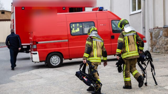 Zwei Feuerwehrleute in Einsatzkleidung mit Helm tragen Atemschutzgeräte zu einem Einsatzfahrzeug.