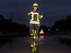 ADAC-Nachttest: Notbremsassistenten erkennen Menschen in genormter Warnschutzkleidung oft nicht Feuerwehrmann in Schutzkleidung mit Reflexstreifen steht nachts in einer Pfütze, daneben ein Warndreieck
