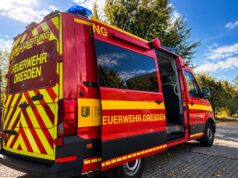 Dresden-Leuben: 83-Jähriger in Waschanlage eingeklemmt – Gabelstapler hebt BMW an Rot-gelber Einsatzleitwagen der Feuerwehr Dresden mit Blaulicht und geöffneter Schiebetür auf einem gepflasterten Platz unter blauem Himmel.