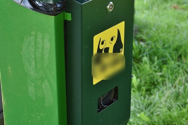 Grüner Hundekotbeutel-Spender mit gelbem Hunde-Aufkleber und Entnahmeschlitz, daneben ein Abfallbehälter, im Hintergrund Rasen.