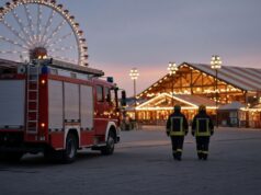 Wiesn 2026: Feuerwehr München bilanziert ruhigen Betrieb – schwerer Betriebsunfall am Schlusstag KI-generiertes Bild: rotes Löschfahrzeug auf einem gepflasterten Festplatz, dahinter ein beleuchtetes Riesenrad und ein Festzelt in der Dämmerung, zwei Feuerwehrleute von hinten.